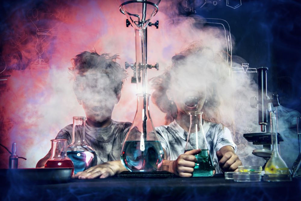 Deux enfants sont assis à une table, devant eux des bocaux de laboratoire munis de tubes et remplis de liquides colorés. Partout, ils se couvrent le visage