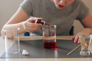 Ein Kind an einem Tisch, vor sich Bechergläser, füllt rote Lebensmittelfarbe in ein Becherglas, in dem sich schon eine Flüssigeit befindet