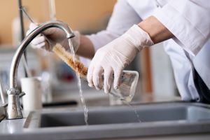 Un robinet dans un laboratoire, deux mains avec des gants et des manches de blouse nettoyant un erlenmeyer avec une brosse sous l'eau courante.