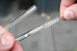 One hand holds two glass straws from the left into the picture, the other hand, from the right into the picture, cleans one of the straws with a small brush provided for this purpose