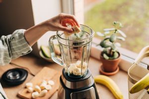 Ausschnitt einer Küche von schräg oben. Ein Standmixer, in den eine Hand Bananenstücken wirft, weitere Bananen im Hintergrund